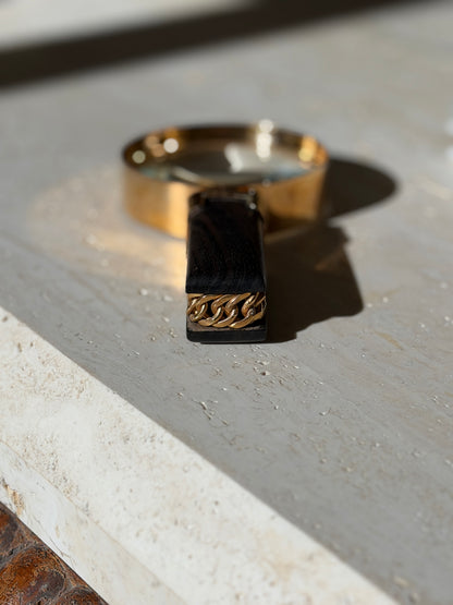 End view of the ebony handle showing the gold chain links set between two slabs of wood, the gilt lens blurred behind