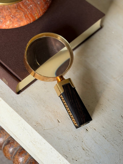 Hermes magnifying glass resting on the corner of a brown clothbound book, the gilt lens rim over the book edge and the Macassar ebony handle with gold chain link inlay extending onto travertine, a red stone ashtray above