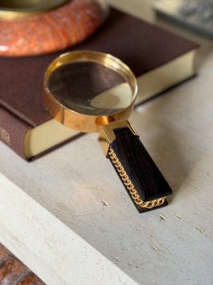 Low angle view of the Hermes magnifier on travertine, the curb chain running between two slabs of Macassar ebony beneath the squared gilt collar, a red stone ashtray softly blurred behind