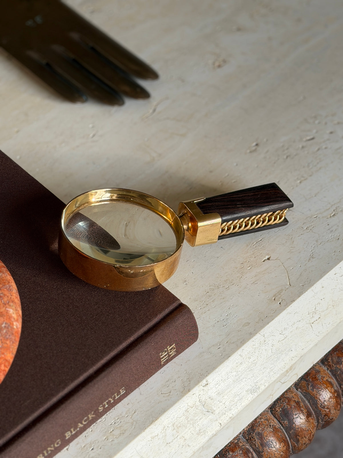 Overhead view of the Hermes gilt brass magnifying glass on the Superfine Tailoring Black Style book, a brass glove form and a bobbin turned table apron framing the scene