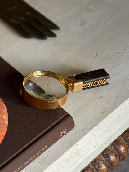 Overhead view of the Hermes gilt brass magnifying glass on the Superfine Tailoring Black Style book, a brass glove form and a bobbin turned table apron framing the scene