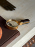 Overhead view of the Hermes gilt brass magnifying glass on the Superfine Tailoring Black Style book, a brass glove form and a bobbin turned table apron framing the scene