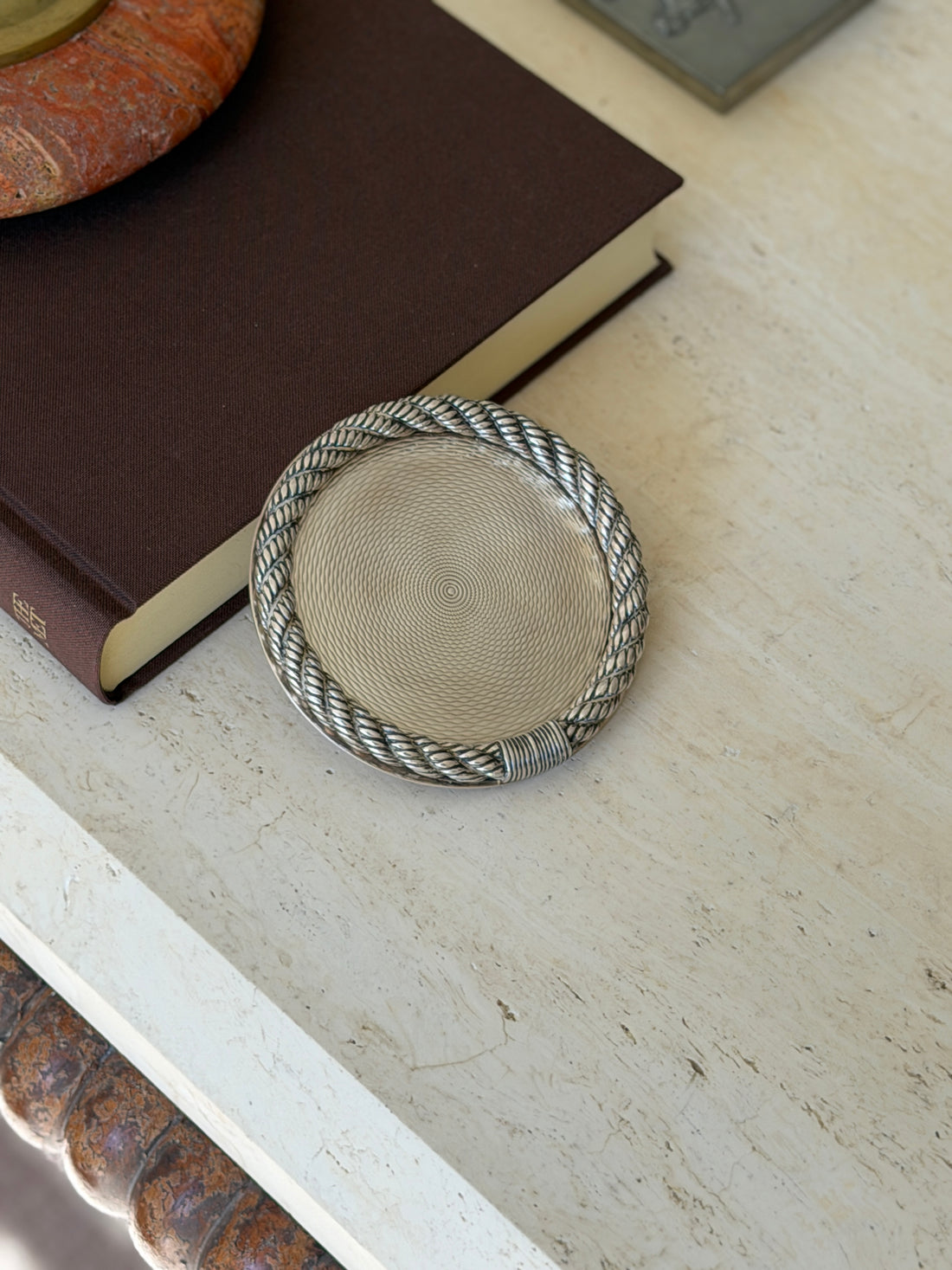 Hermes cordage coaster resting against a brown book, the oxidized rope twist framing the radiating guilloche center
