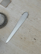 Hermes silver-tone letter opener with a sculptural spur handle, the long tapered blade lying diagonally on travertine beside a silver chain-rimmed coaster