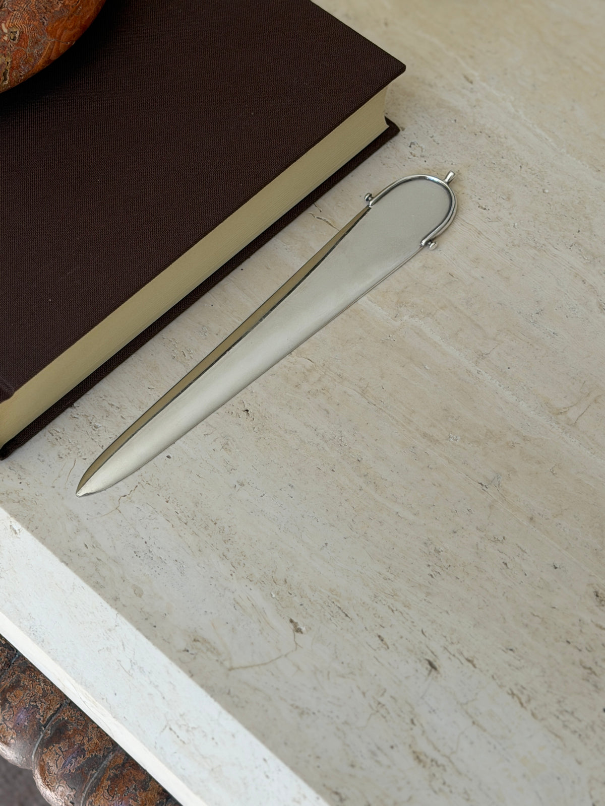 Hermes letter opener lying on a travertine table alongside a brown book, the polished blade tapering to a fine point