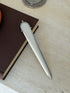 Hermes spur handle letter opener angled against the corner of a brown book, light catching the smooth rounded handle and blade edge