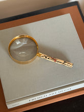 Hermes gilt magnifying glass with a cream Japanese Meiji period handle inlaid with flowering vines and birds, lying on a gray linen book titled French Art Deco