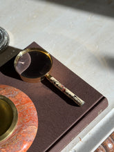 High angle view of the gilt magnifier on a brown book, sunlight across the lens and the softly worn cream handle