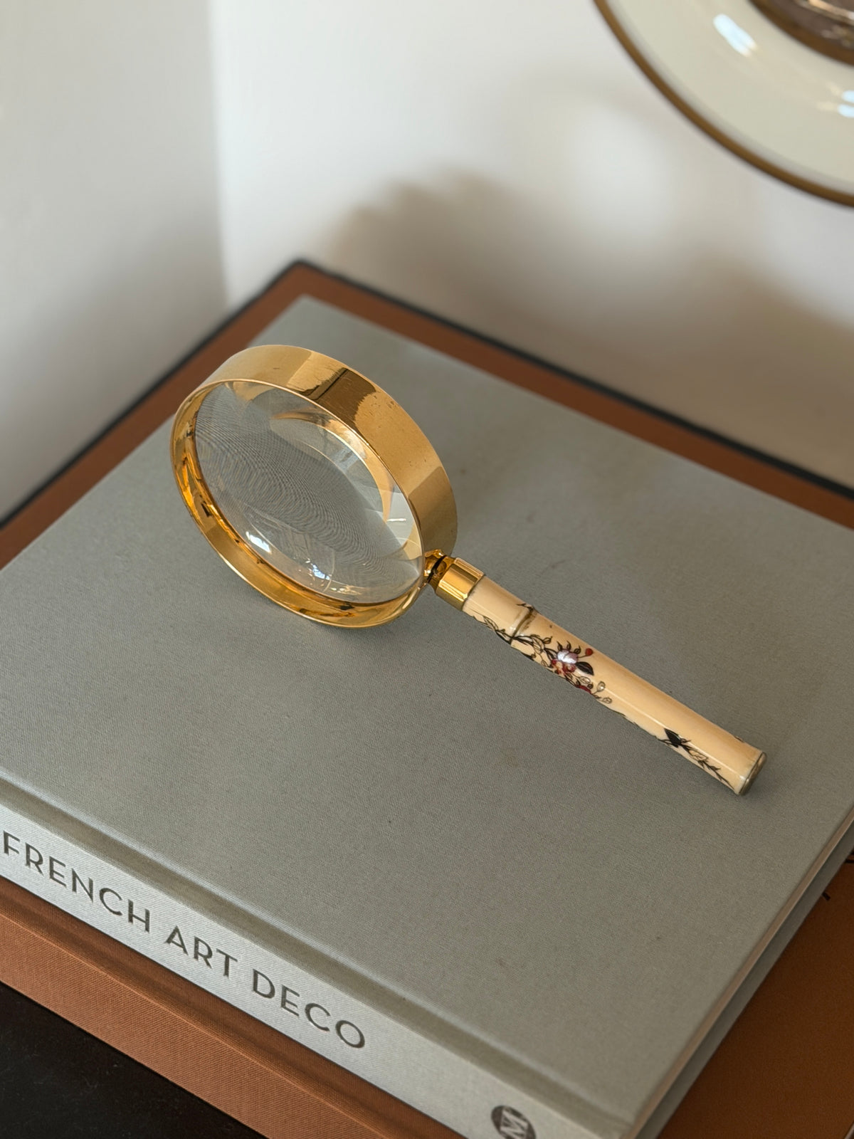 Hermes magnifier resting at an angle on a gray book, the deep polished gilt rim catching light above the inlaid cream handle