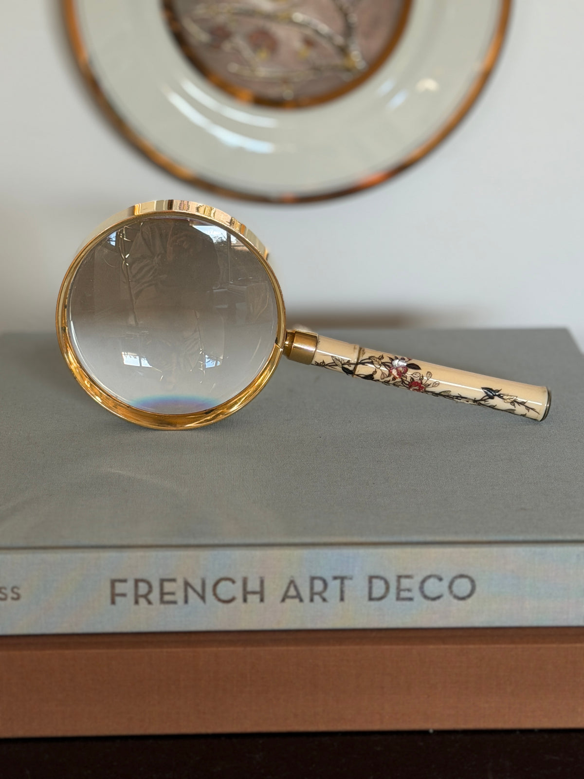Front view of the Hermes magnifying glass standing on a stack of books, a gilt rimmed porcelain plate in the background