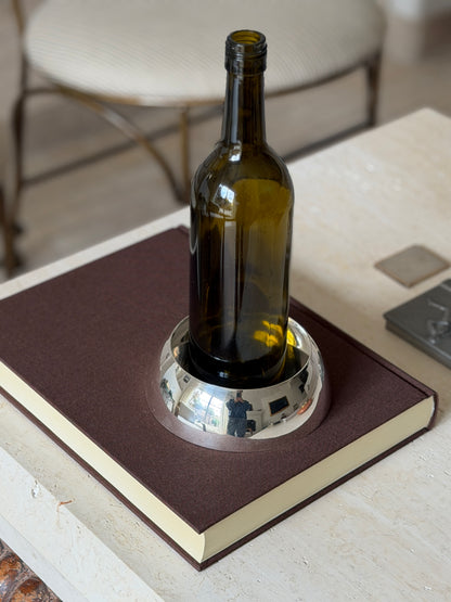 Hermes Paris silver bottle coaster holding a green wine bottle on a brown book, its domed mirror finish reflecting the room