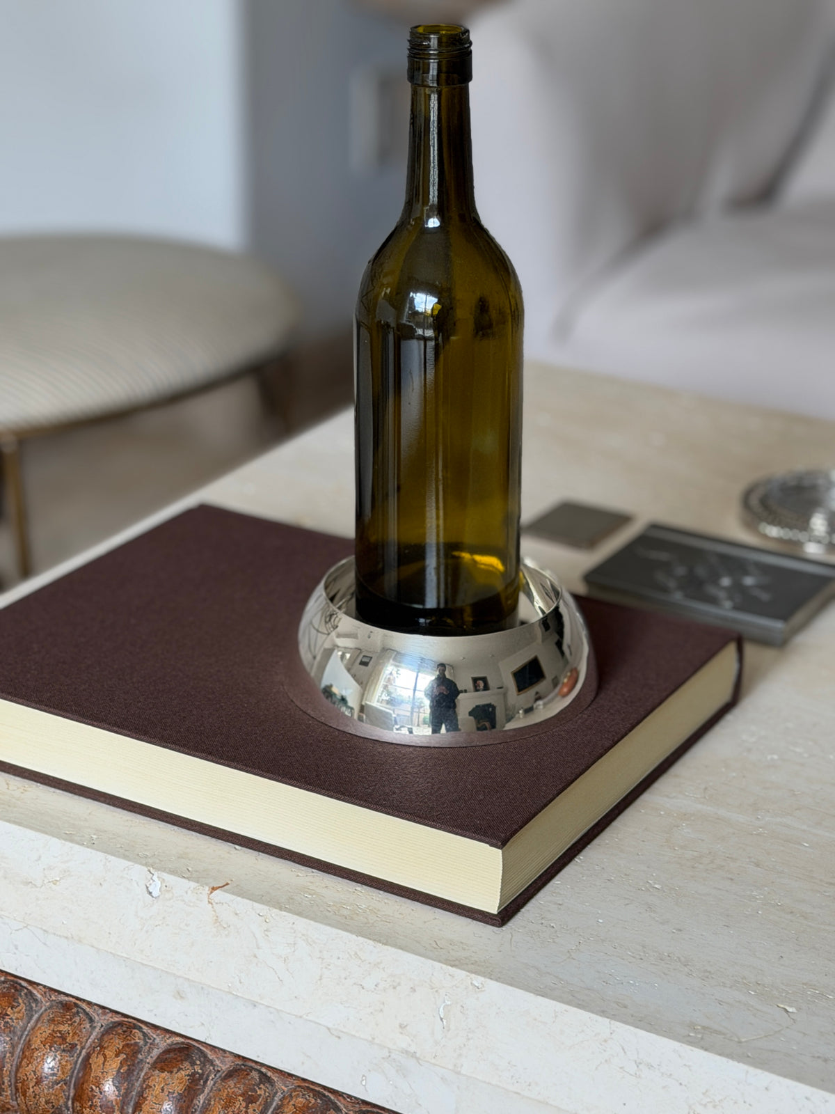 Wine bottle set in the polished Hermes coaster on a brown book atop travertine, a striped chair and sofa softly blurred behind