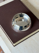 Empty Hermes bottle coaster on a brown clothbound book, the rounded silver wall and plain polished base in view
