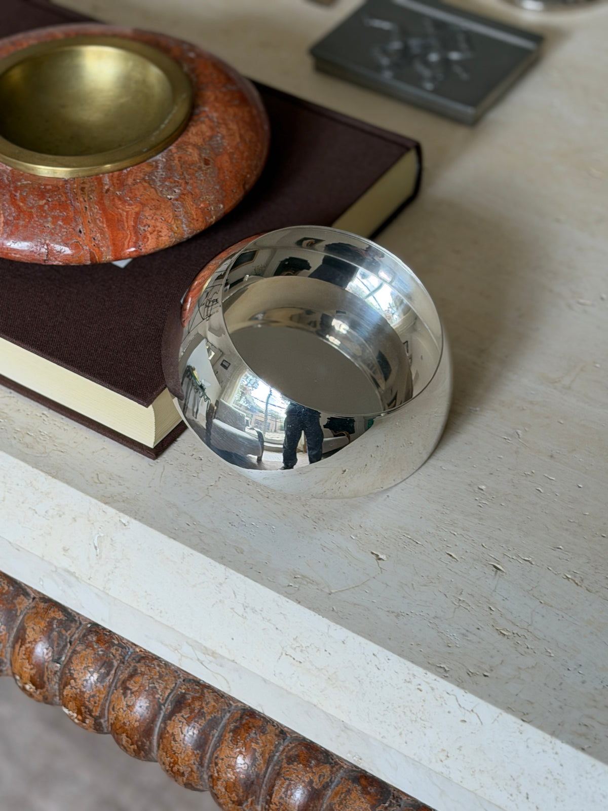 Hermes silver bottle coaster resting on travertine beside a red stone ashtray with brass well on a brown book
