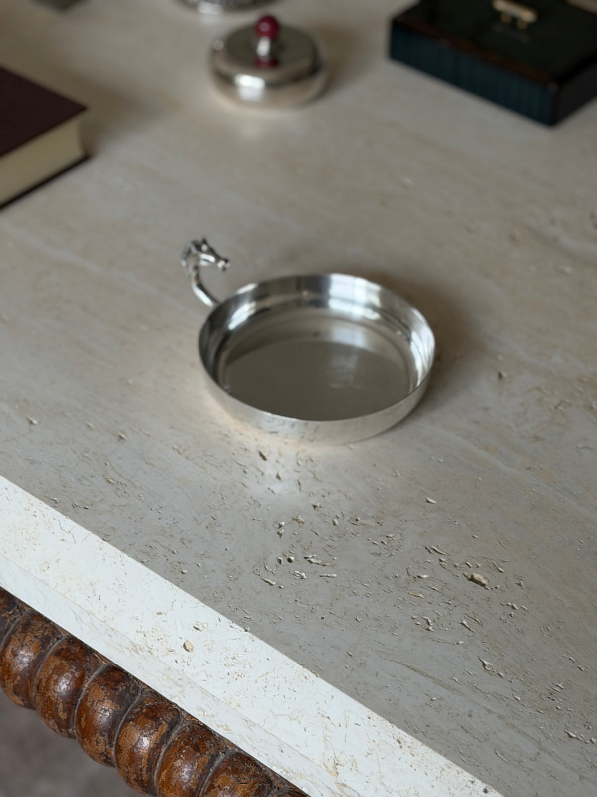 Round silver plated dish near the travertine table edge and its bobbin turned wood apron, a lidded box and dark box blurred behind