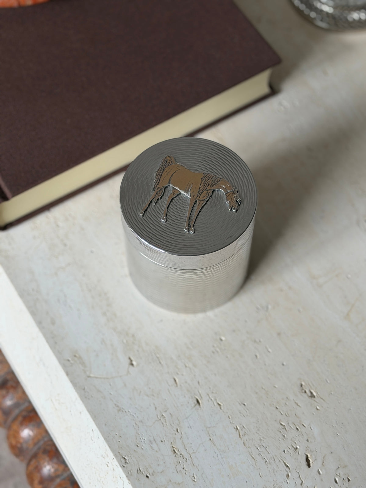 Hermes solid silver lidded box with a gilt horse on the lid, its body engine turned with fine wavy lines, on travertine beside a brown book