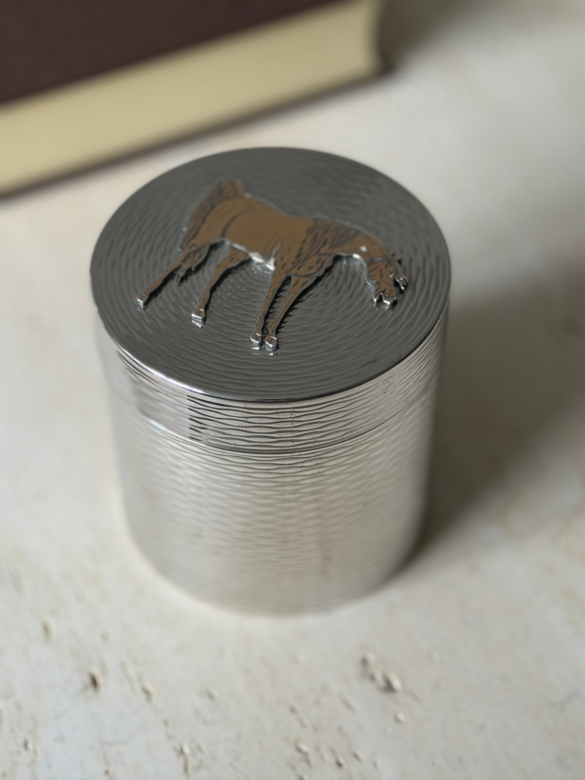Hermes cylindrical silver box on a travertine console with a brown book, a red stone ashtray, and a silver coaster behind