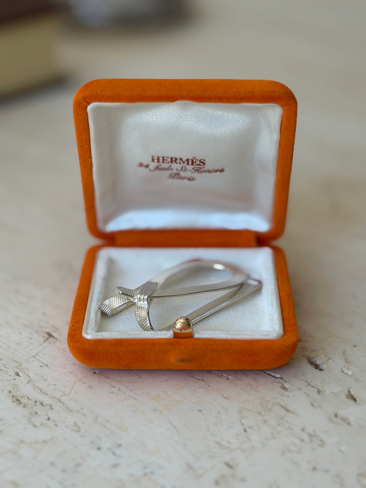 Hermes sterling silver money clip in its open orange suede box, a textured strap crossing the loop above the Paris address lining