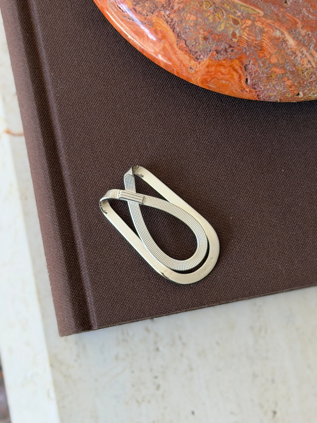Crossed strap money clip on a brown book, pairing a polished outer loop with a diamond pattern strap and fluted band