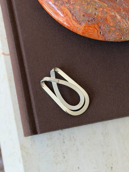Crossed strap money clip on a brown book, pairing a polished outer loop with a diamond pattern strap and fluted band