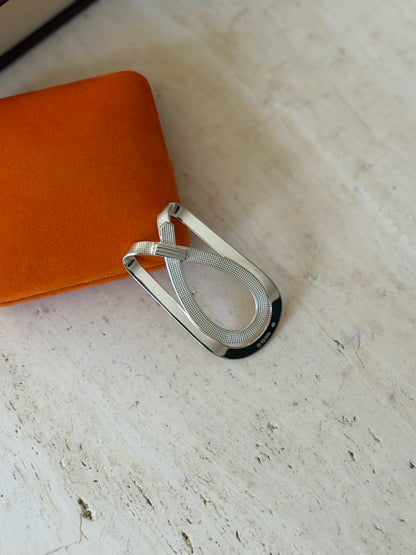 Hermes crossed strap money clip resting against the orange suede box, sun catching the textured strap and polished silver