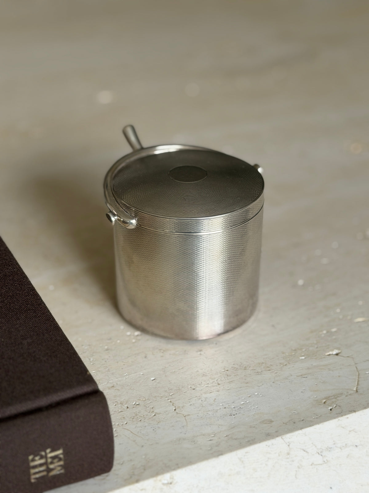 Hermes sterling silver cylindrical cigarette box with engine turned guilloche, a spur shaped handle arching over the lid, beside a brown book on travertine