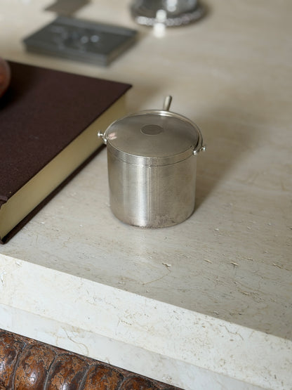 Cylindrical silver box on a travertine table edge near a brown book, the fine engine turned body and swinging spur handle on button pivots