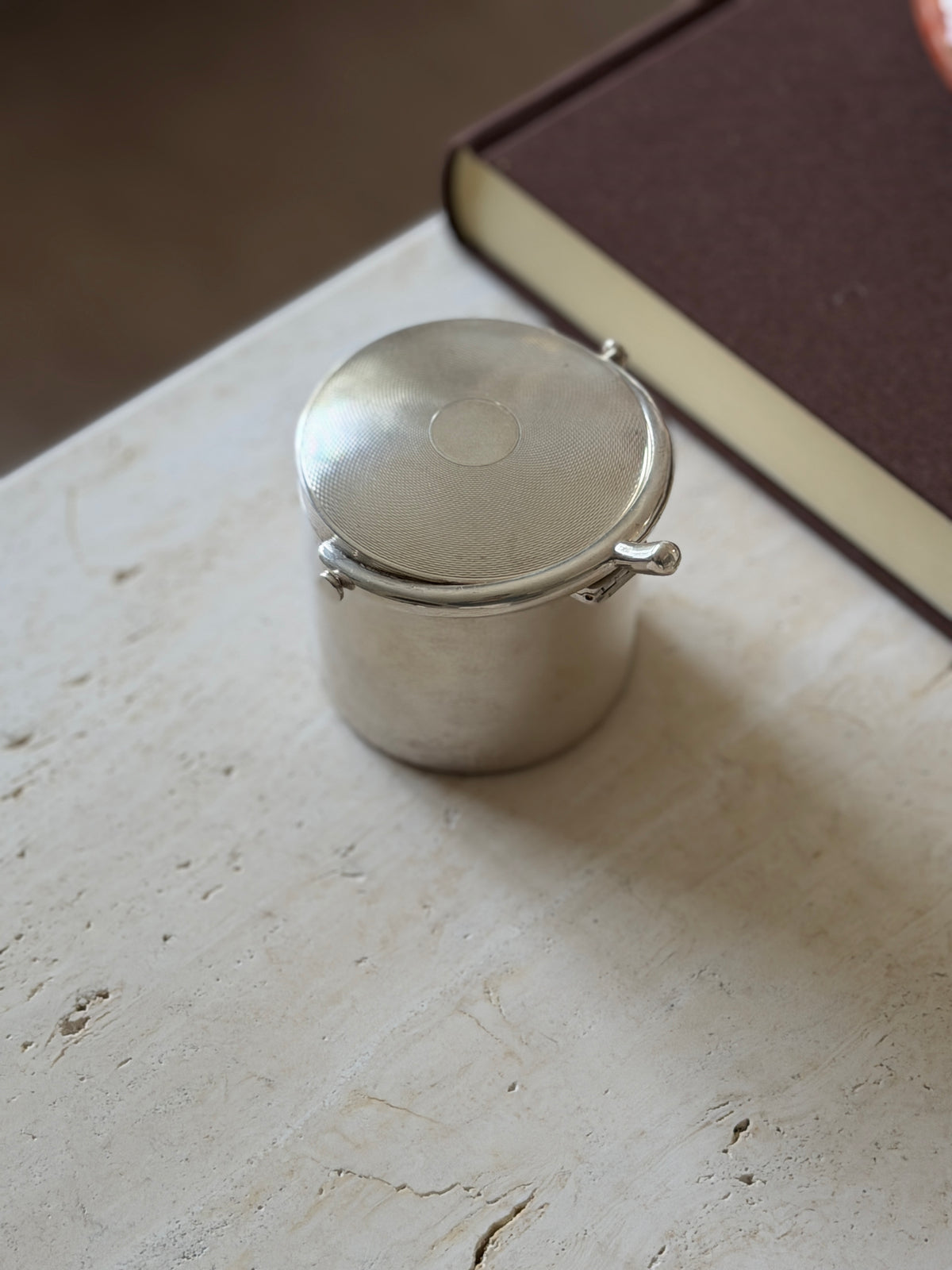 Angled view of the cylindrical cigarette box near a brown book, sunlight across the engine turned lid and the polished spur handle