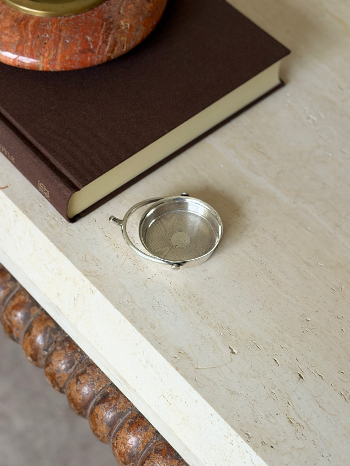 Small sterling silver spur ashtray on a travertine table beside a brown book topped with a red stone ashtray