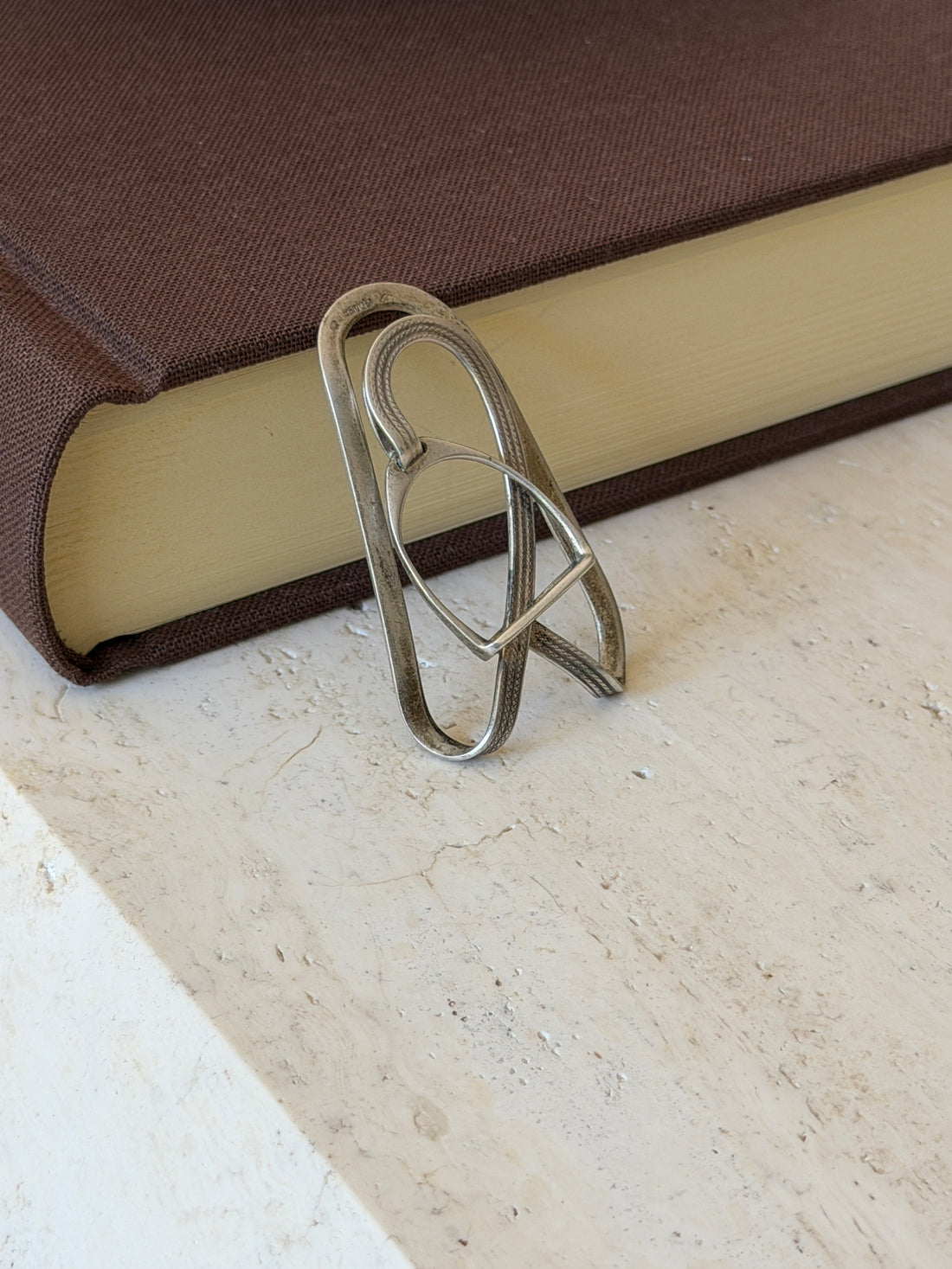 Stirrup money clip standing against a brown book on travertine, the worn patina showing along the plain outer loop