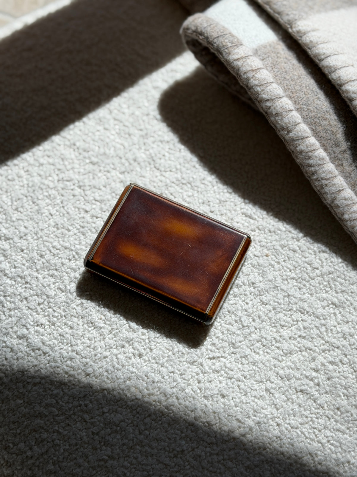 Keller Paris lacquer box on a cream boucle surface in raking sunlight, light surface wear visible on the polished lid