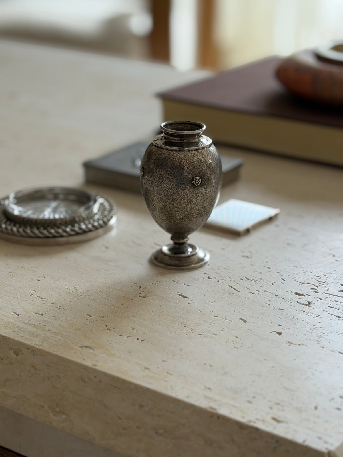 Textured silver ovoid vase with a flared rim and stepped collar on a travertine table, a brown book and red stone ashtray blurred behind