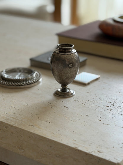 Textured silver ovoid vase with a flared rim and stepped collar on a travertine table, a brown book and red stone ashtray blurred behind