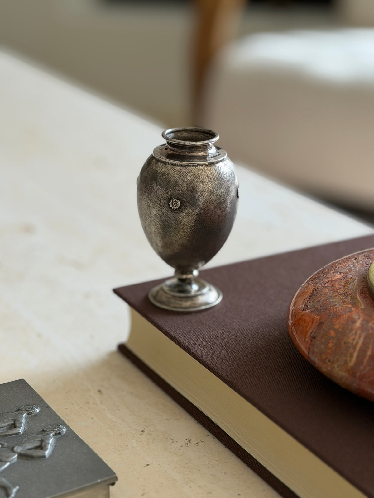 Silver bud vase with floral rosettes on the corner of a brown book next to a red stone ashtray, a pewter box in the foreground