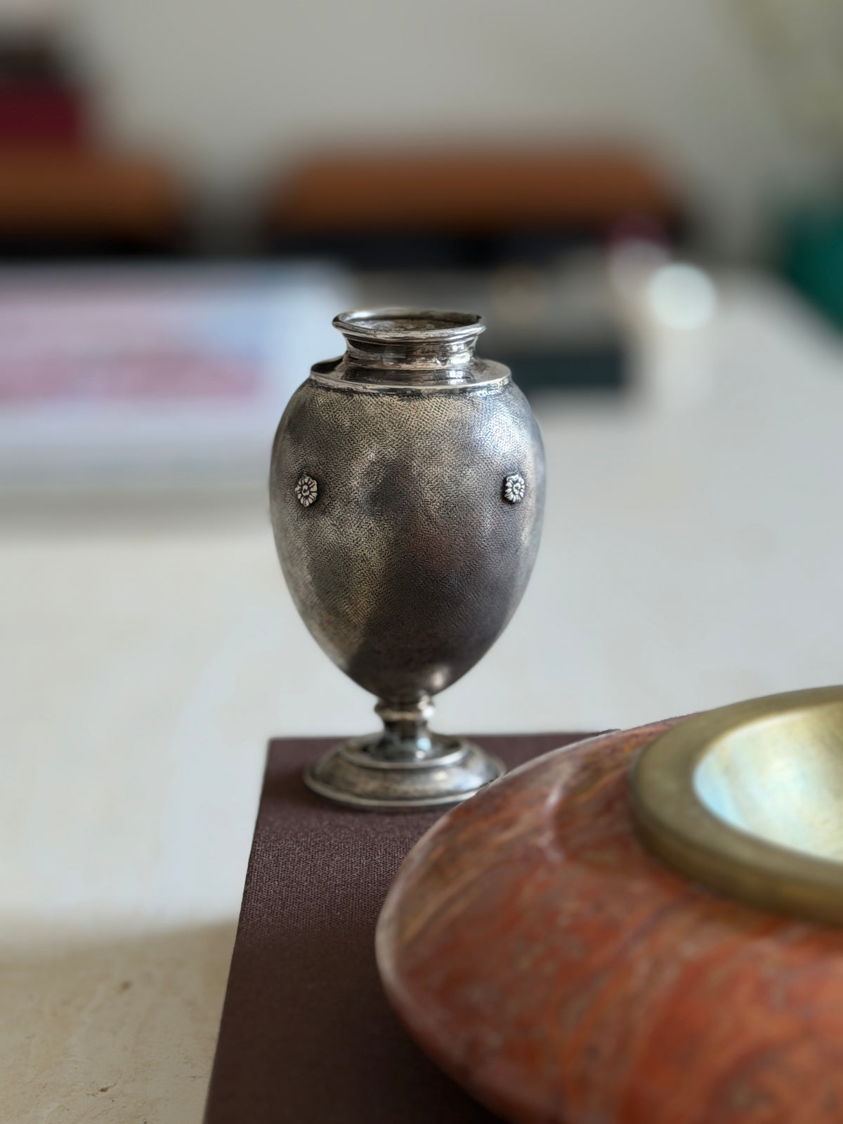 Buccellati textured silver vase on a brown book edge, two rosettes framing the body, the red stone ashtray in soft focus