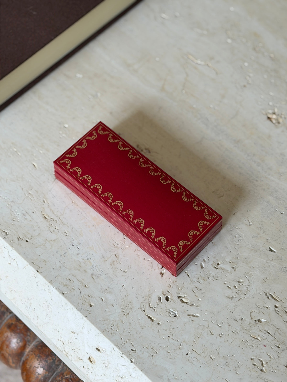 Closed red Cartier presentation case with gold garland border on travertine, a bobbin turned table apron below