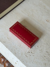 Closed red Cartier presentation case with gold garland border on travertine, a bobbin turned table apron below