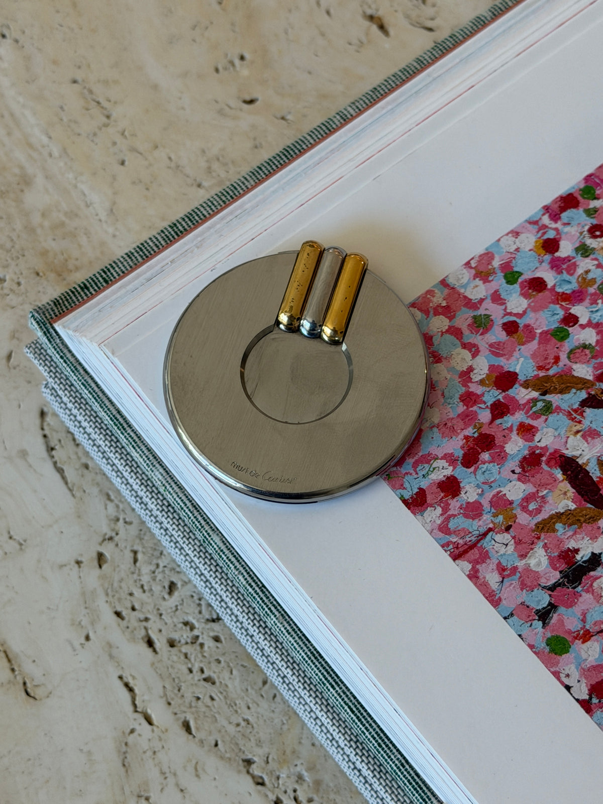 Overhead view of the closed Cartier cigar cutter on a white page beside a colorful painted plate in an art book