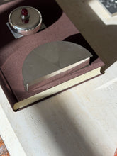 Olivetti half moon box in engine turned silver plate on a brown clothbound book, the lid divided into three textured sectors, a lidded silver box behind