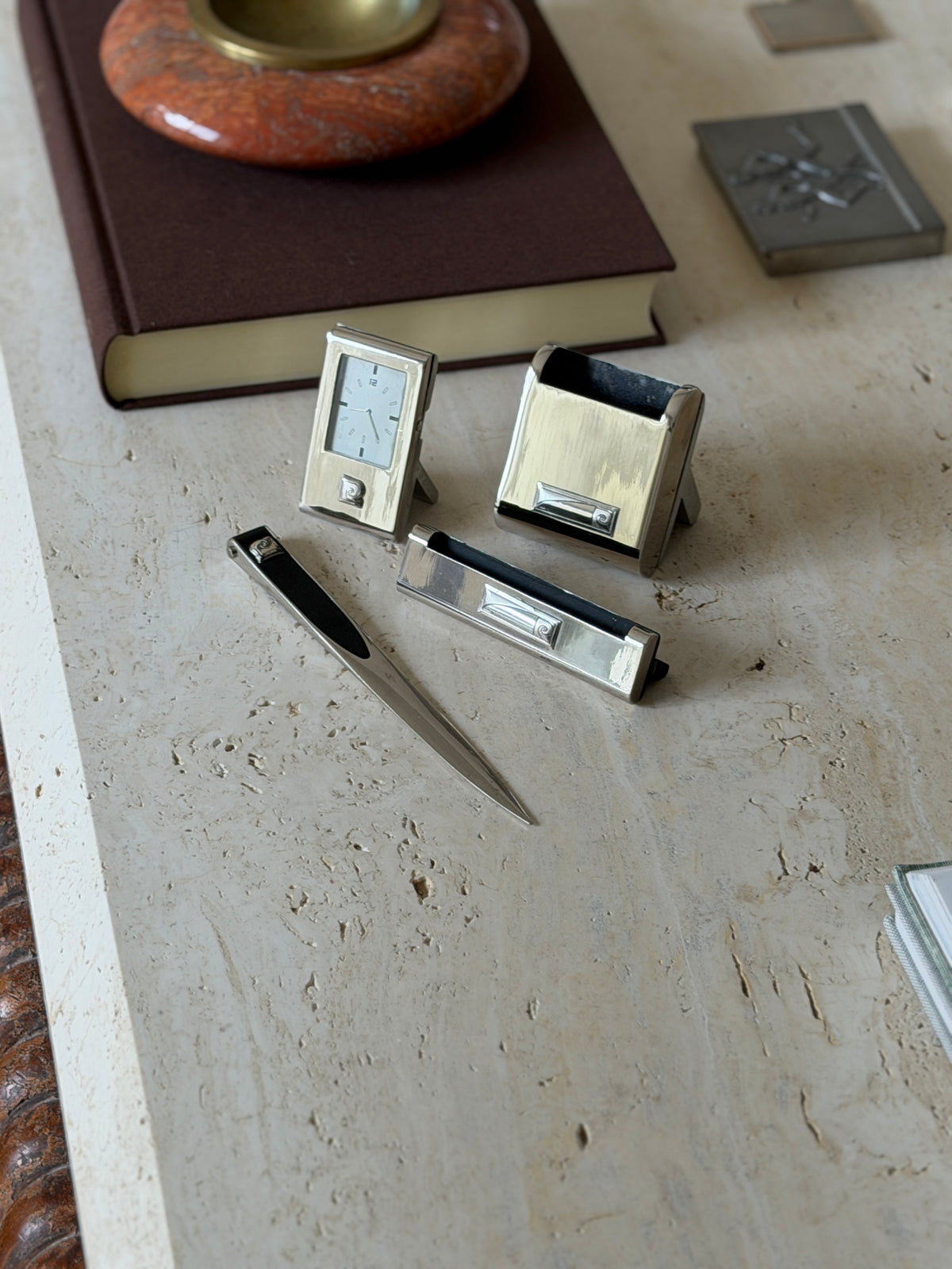 Pierre Cardin chrome desk set on travertine, clock, pen holder, card holder and letter opener each with the spiral logo plaque, a red stone ashtray behind