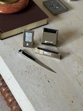 Pierre Cardin chrome desk clock, holders and letter opener arranged on travertine beside a brown book and pewter box