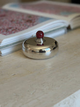 Overhead view of the Puiforcat candy dish on a travertine table near an open art book, the red finial centered on the lid