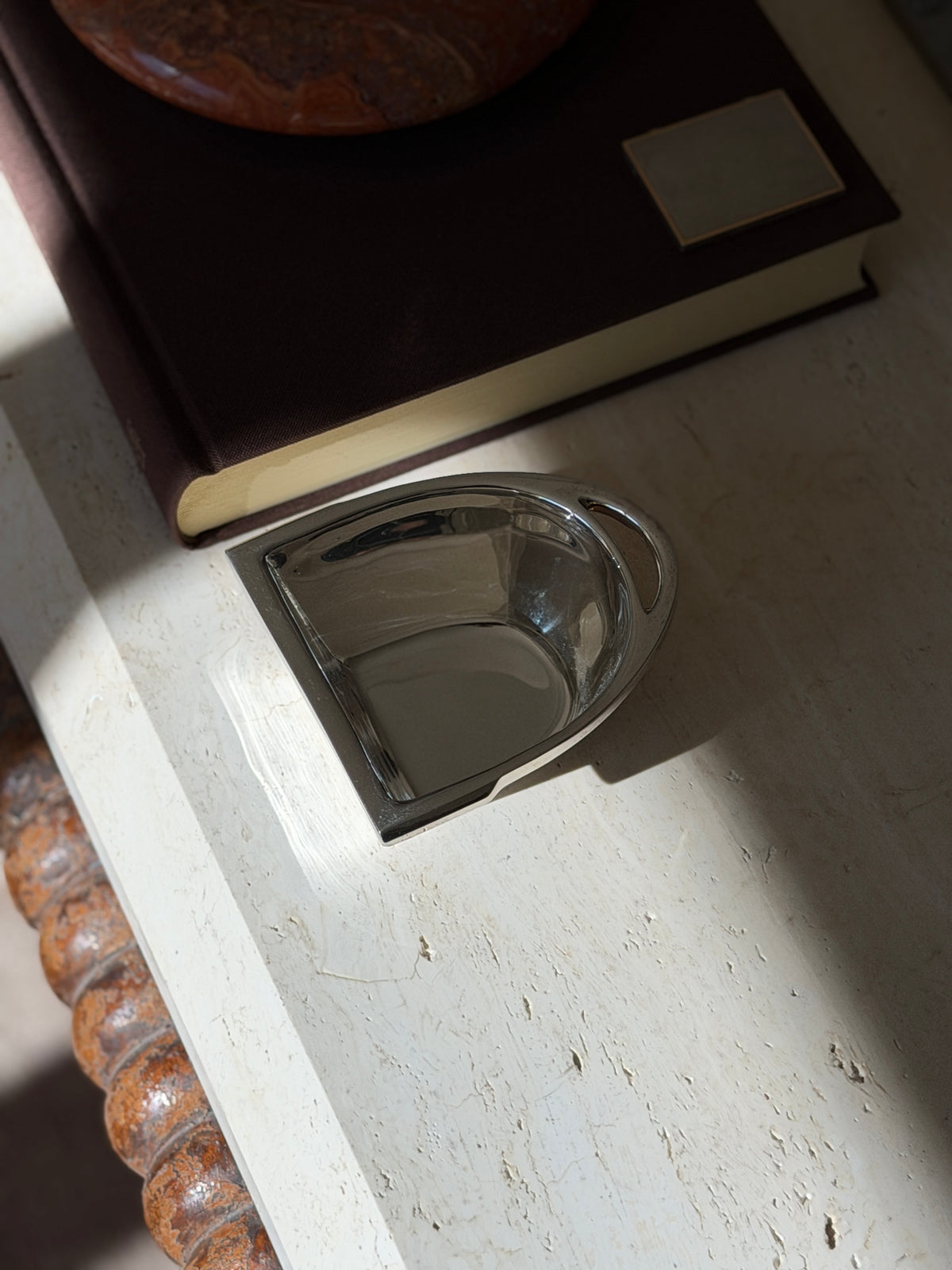 Ralph Lauren stirrup bowl on travertine beneath a brown book and red stone ashtray, the polished silver reflecting the window light