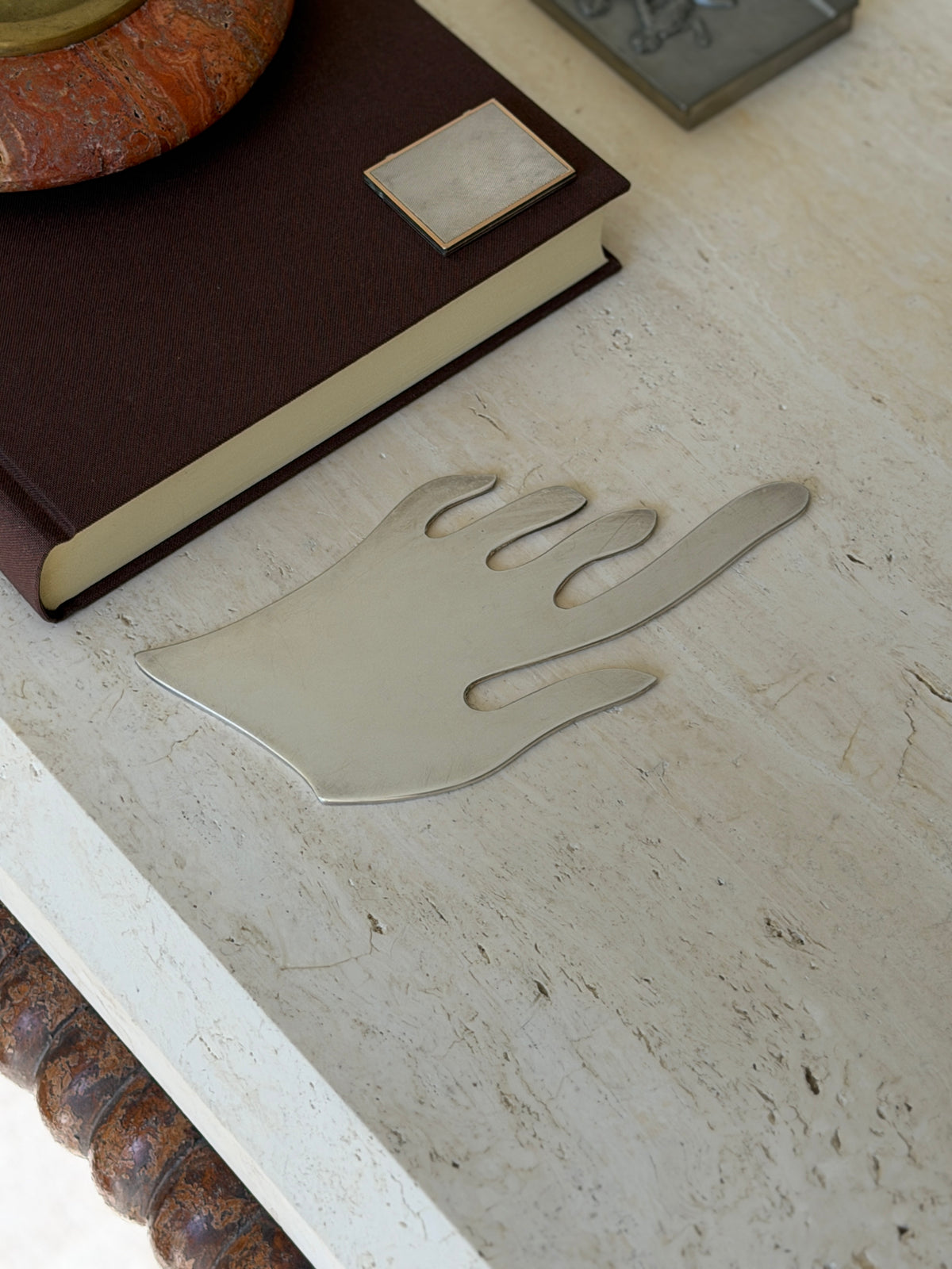 Minimalist cutout hand lying flat on travertine, the long pointing finger and flame like digits in brushed silver