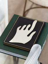 Silver hand sculpture on a black and gold bordered book atop an Audemars Piguet volume, a frosted glass object in the foreground