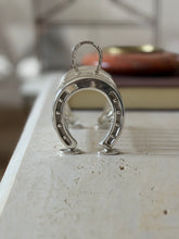 Front view of the equestrian toast rack, the studded horseshoe end standing on round pad feet with a red stone ashtray blurred behind