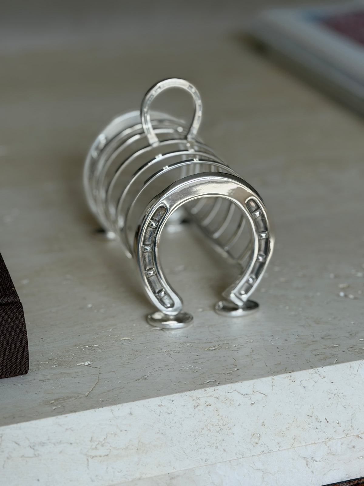 Horseshoe toast rack on travertine beside a brown book, the polished studded shoe and arched dividers in sharp focus