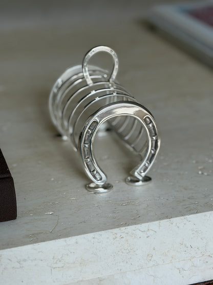 Horseshoe toast rack on travertine beside a brown book, the polished studded shoe and arched dividers in sharp focus