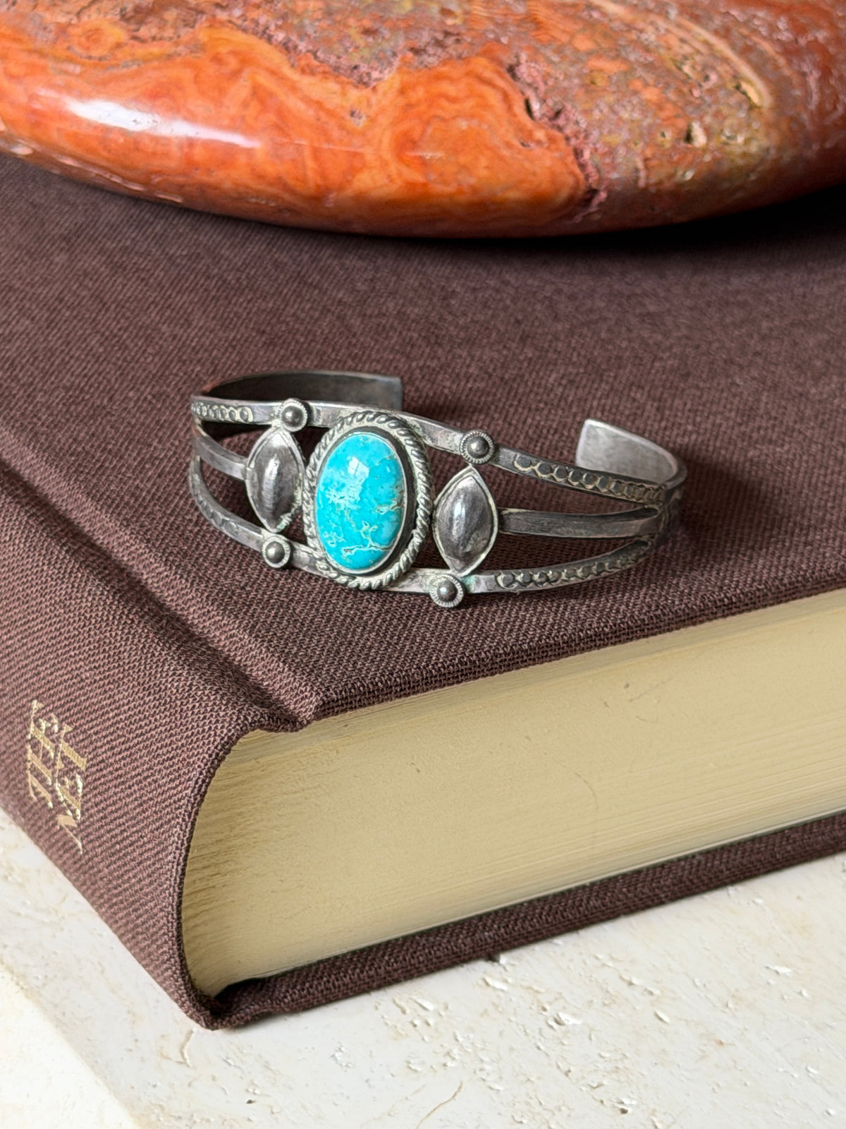 Sterling turquoise cuff resting on a brown clothbound book beneath a polished red stone, the oxidized patina visible along the bands