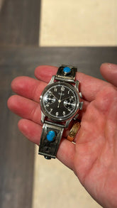 Front view of 1940s Heuer black dial chronograph on stamped sterling silver turquoise bracelet held in palm